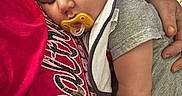 Carson is registered to the contest to win money with this photo: baby, sleeping, pacifier, person, arm, clothing, bib, red_shirt, text_on_clothing, quilt, blanket, comfort, indoor, close_up, portrait, skin, hand, relaxation, cute, toddler