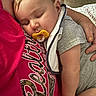 baby, sleeping, pacifier, person, arm, clothing, bib, red_shirt, text_on_clothing, quilt, blanket, comfort, indoor, close_up, portrait, skin, hand, relaxation, cute, toddler