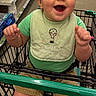 Carson is registered to the contest to win money with this photo: baby, cart, child, cute, footwear, green_bib, green_shirt, hands_raised, happy, indoor, infant, person, products, retail, shelves, shopping, shopping_cart, shorts, smiling, store_aisle