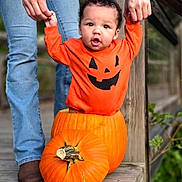 Zakai is registered to the contest to win money with this photo: adult_hand, baby, boots, child, costume, cute, fall, festive, greenery, halloween, holding_hands, holiday, jeans, orange_clothing, outdoor, portrait, pumpkin, seasonal, smiling, wooden_deck