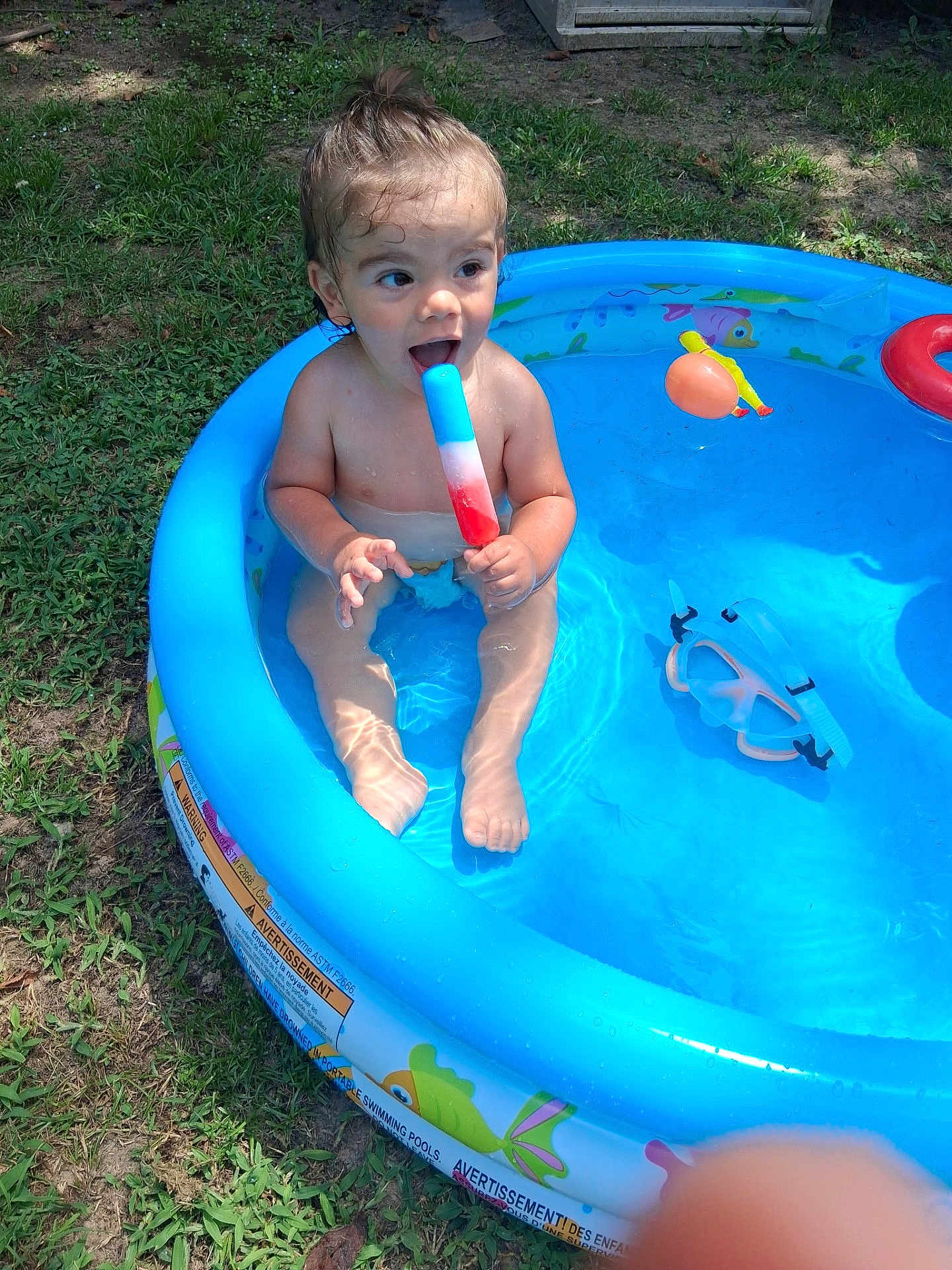 Xatthias is registered to the contest to win money with this photo: blue, child, colorful, fun, goggles, grass, happy, inflatable, outdoor, playing, pool, popsicle, sitting, skin, summer, sunlight, toddler, toy, water, wet
