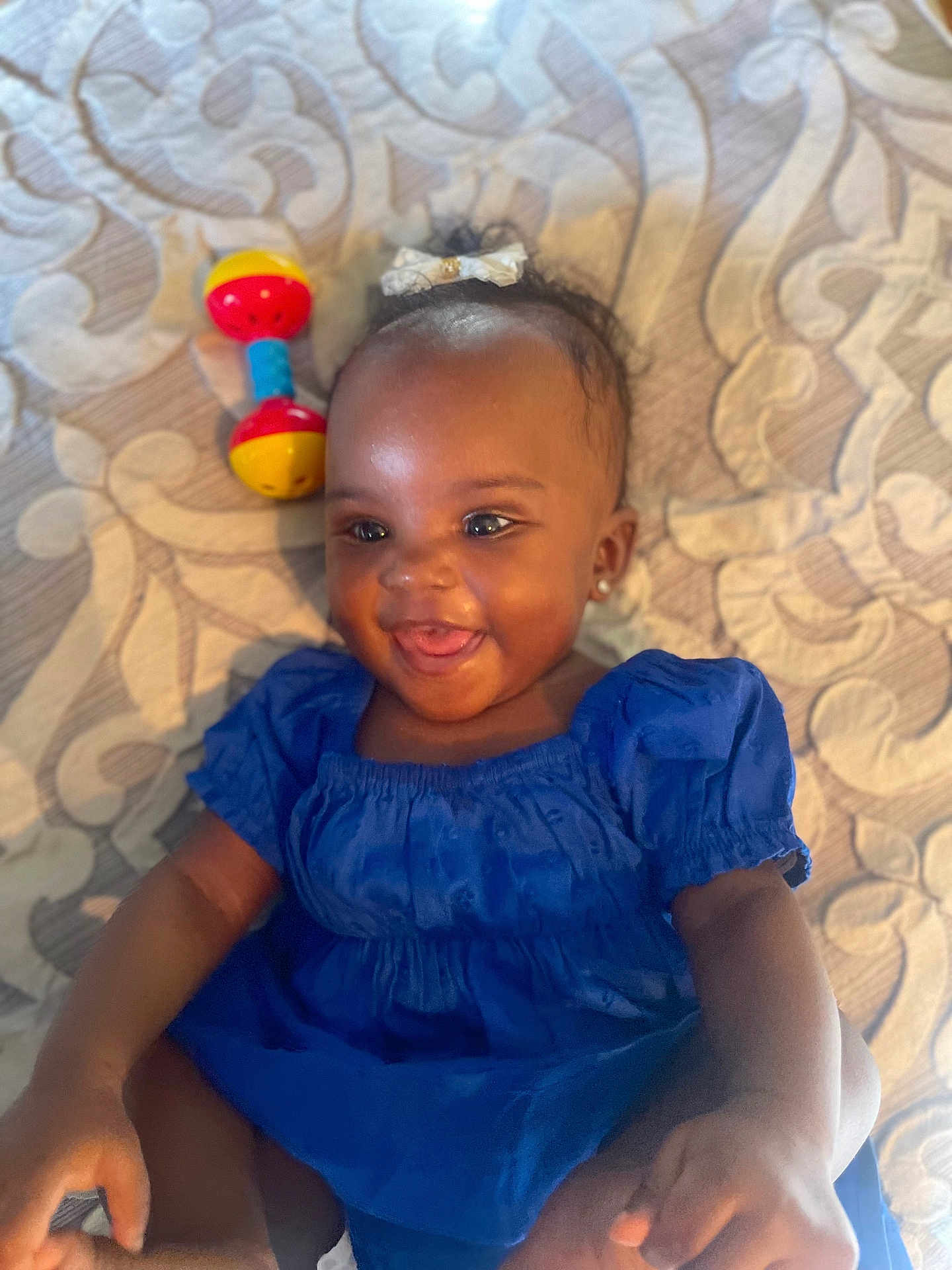Kaija is registered to the contest to win money with this photo: baby, child, blue_dress, toy, rattle, smiling, lying_down, earrings, bow, patterned_quilt, infant, happy, cute, skin, hand, foot, indoors, portrait, closeup, person