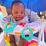 baby, smiling, happy, infant, baby_seat, toys, umbrella, outdoor, colorful, playful, child, sunny_day, clothing, pacifier, rainbow_toy, plastic_toys, face, person, seat, cute