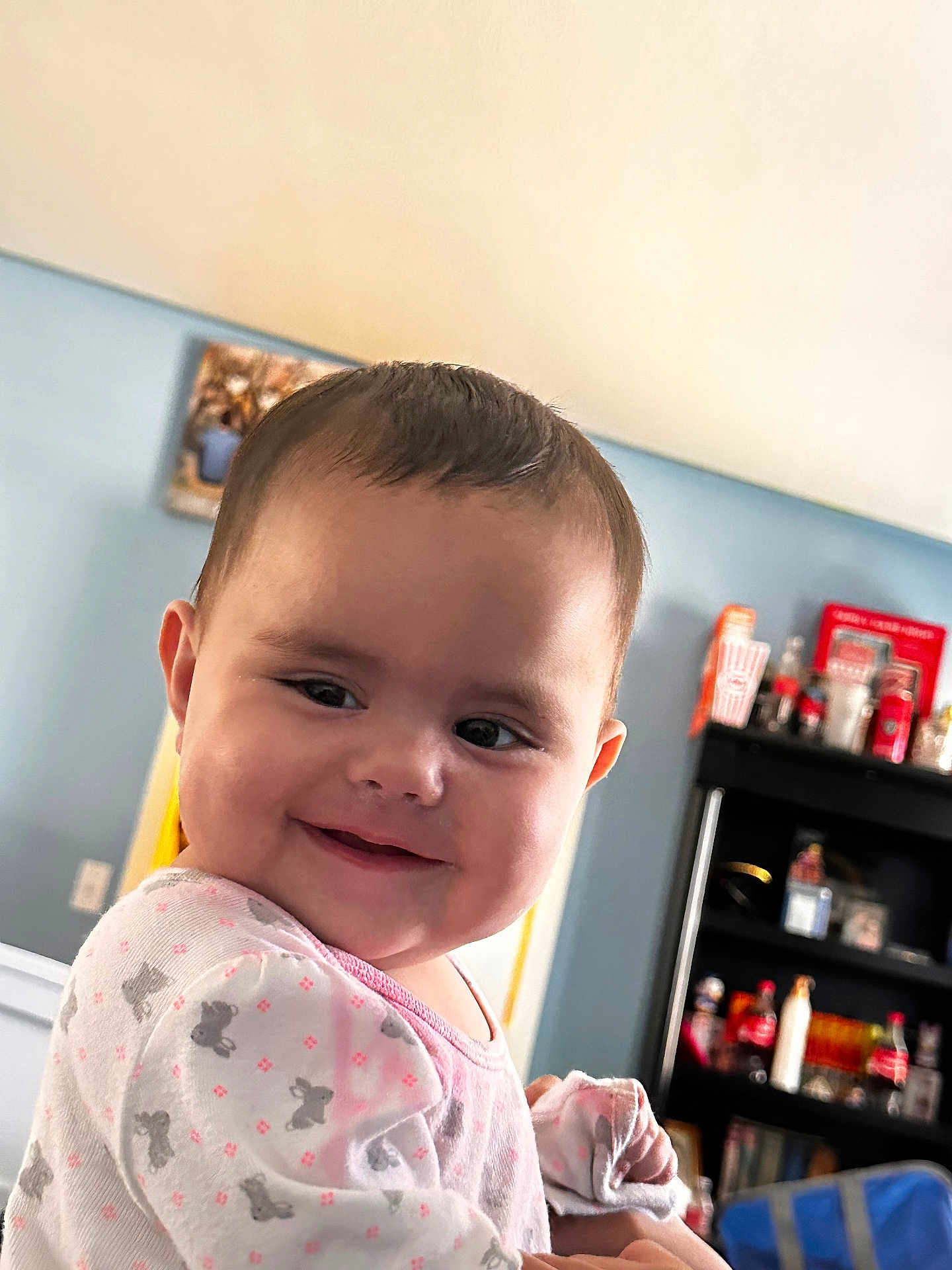 Ellie is registered to the contest to win money with this photo: baby, child, smile, indoor, onesie, face, person, cute, happy, sitting, furniture, shelf, bottle, toy, clothing, head, portrait, home, expression, infant