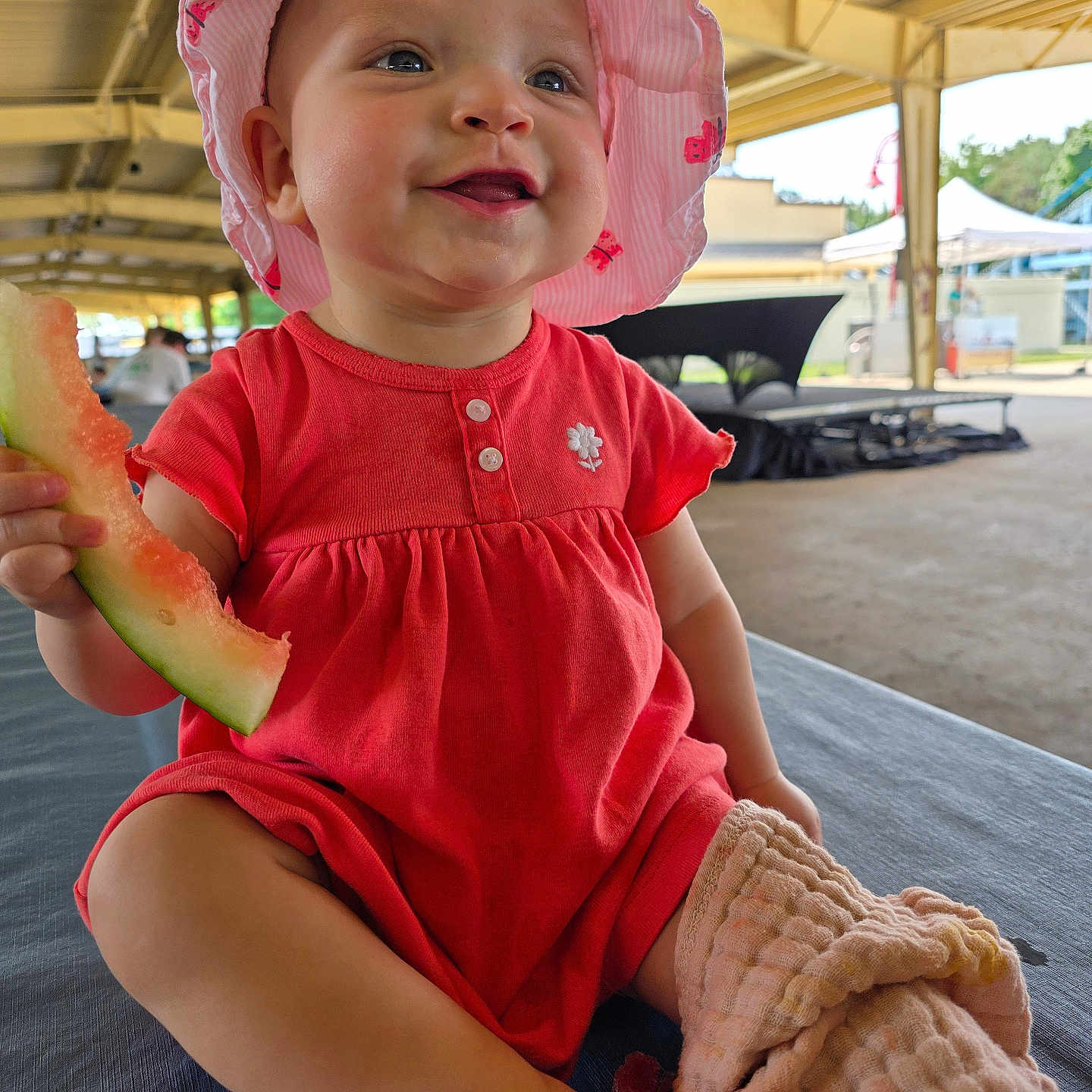 Elowyn is registered to the contest to win money with this photo: baby, child, pink_hat, pink_outfit, watermelon, food, outdoor, pavilion, blanket, smiling, happy, sitting, infant, summer, sunlight, cute, portrait, headwear, clothing, person