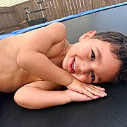 Felipe joined the competition — help win amazing prizes! child, smiling, outdoor, trampoline, backyard, portrait, curly_hair, brown_hair, lying_down, happy, face, teeth, arms, hands, diaper, playground, fence, sunlight, recreation, photography