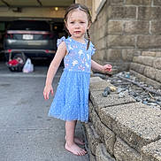 Kenny is registered to the contest to win money with this photo: toddler, child, barefoot, blue_dress, stone_wall, rocks, outdoor, sidewalk, car, baby_carrier, curly_hair, braids, expression, concrete, daylight, person, standing, casual, curious, young_child