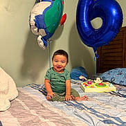 Jonathan is registered to the contest to win money with this photo: toddler, child, balloon, blue_balloon, number_6, toy, bed, bedding, smile, happy, indoor, green_shirt, pants, wooden_headboard, pillow, blanket, cartoon_balloon, celebration, birthday, cute