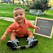 Beckham is registered to the contest to win money with this photo: baby, child, outdoor, grass, greenery, smiling, orange_shirt, sign, milestone, shoes, orthopedic, happy, portrait, sitting, cute, person, nature, daylight, flower_bush, yard