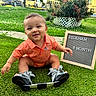 baby, child, outdoor, grass, greenery, smiling, orange_shirt, sign, milestone, shoes, orthopedic, happy, portrait, sitting, cute, person, nature, daylight, flower_bush, yard