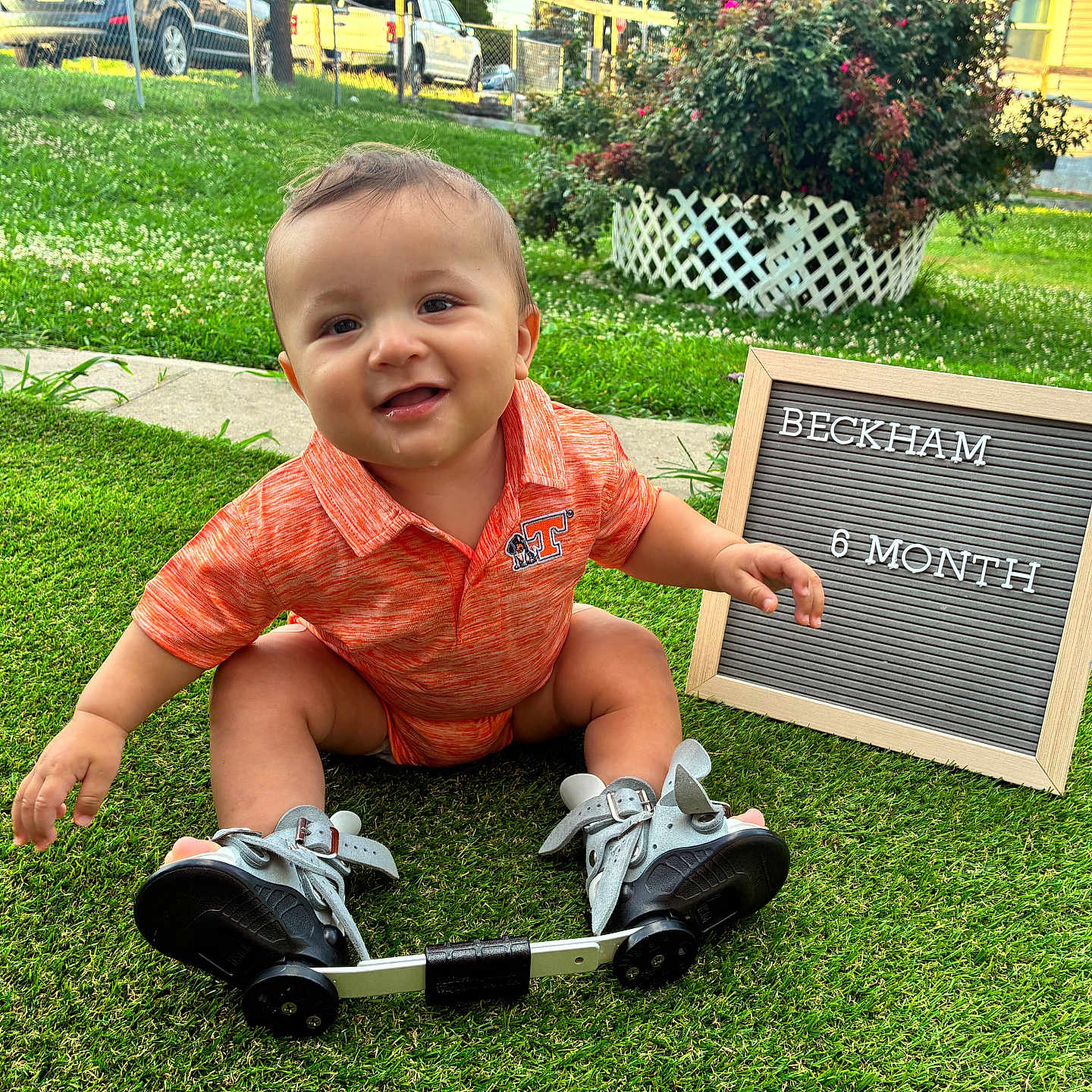 Beckham is registered to the contest to win money with this photo: baby, child, outdoor, grass, greenery, smiling, orange_shirt, sign, milestone, shoes, orthopedic, happy, portrait, sitting, cute, person, nature, daylight, flower_bush, yard