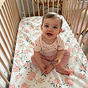 Gabriela participe au concours pour gagner de l'argent avec cette photo : baby, child, crib, wooden_crib, floral_bedding, pink_clothing, smiling, sitting, indoors, cute, happy, person, infant, blue_eyes, soft_lighting, room, floor, chair, foot, hand