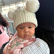 Tru is registered to the contest to win money with this photo: baby, child, face, hat, knit_hat, pom_poms, floral_clothing, person, holding, indoor, window, adult, beanie, black_clothing, blanket, curious, portrait, closeup, warm_clothing, soft_light
