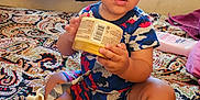 Adrian is registered to the contest to win money with this photo: toddler, child, baby, sitting, holding_object, indoor, bedspread, paisley_pattern, window, curtains, socks, pacifier, container, barcode, expression, hair_tied, room, furniture, toy, clothing