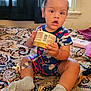 toddler, child, baby, sitting, holding_object, indoor, bedspread, paisley_pattern, window, curtains, socks, pacifier, container, barcode, expression, hair_tied, room, furniture, toy, clothing