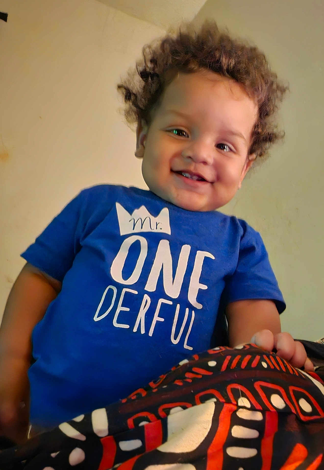 Adrian is registered to the contest to win money with this photo: toddler, child, smiling, curly_hair, blue_shirt, text_on_clothing, indoor, portrait, happy, person, cute, young_child, casual_clothing, patterned_fabric, hand, face, head, skin, human, one_year_old