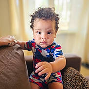 Adrian is registered to the contest to win money with this photo: toddler, child, curly_hair, camo_clothing, indoor, couch, hand, face, sitting, portrait, furniture, window, natural_light, skin, expression, person, home, leopard_print, focus, baby