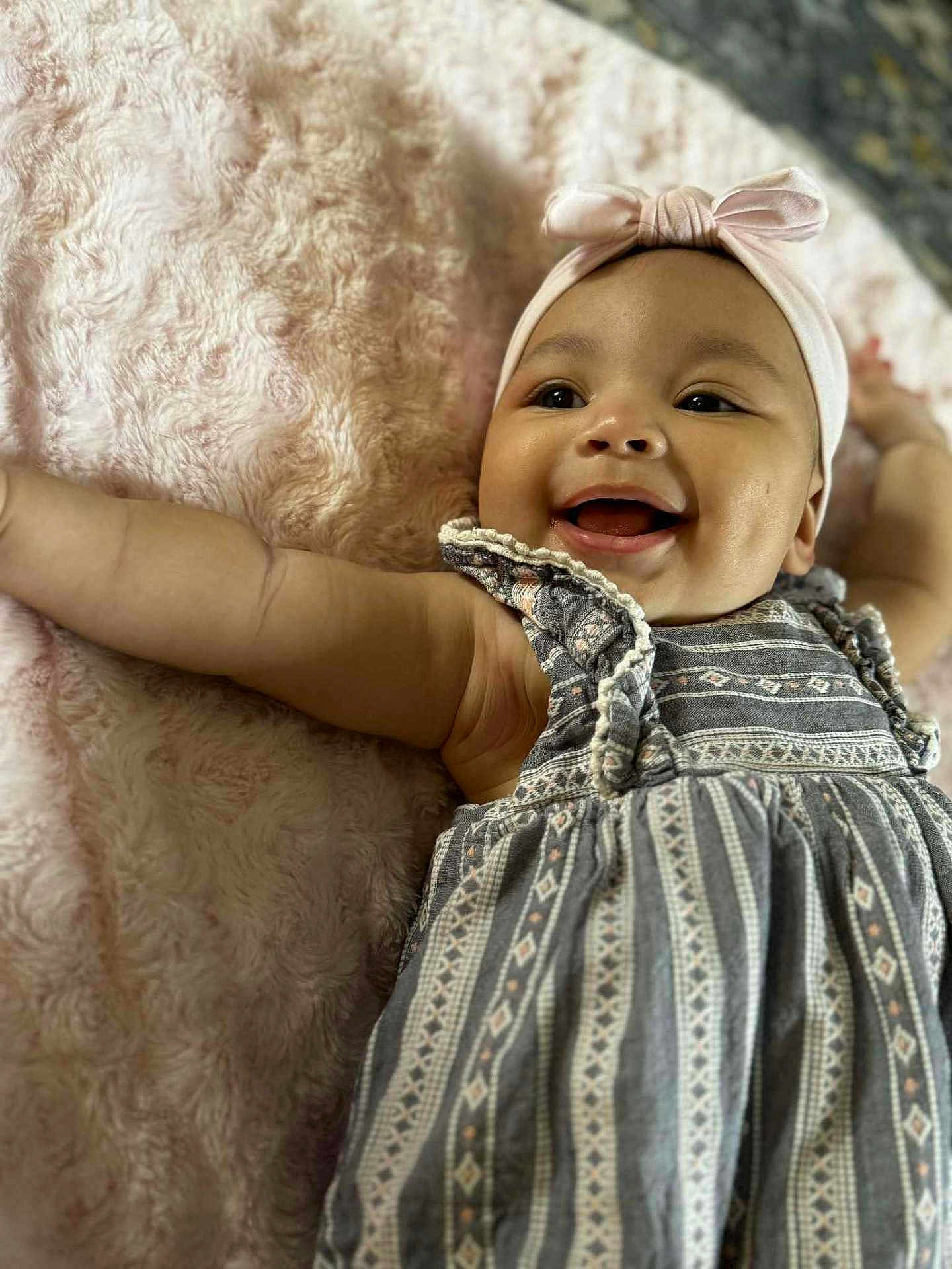 Isabella joined the competition — help win amazing prizes! baby, smiling, headband, dress, infant, happy, cute, child, portrait, lying_down, skin, face, arm, blanket, textile, patterned_clothing, indoors, closeup, person, cheerful
