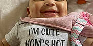 Isabella is registered to the contest to win money with this photo: baby, smiling, onesie, text_on_clothing, toy, blanket, infant, cute, happy, face, child, pink, white, clothing, lying_down, cheeks, indoor, playful, colorful, bib
