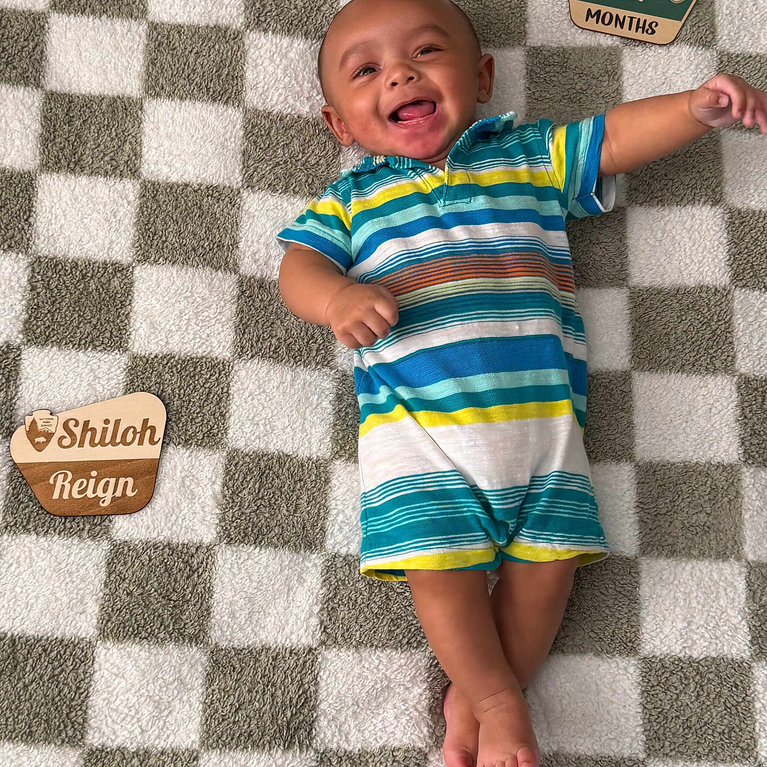 Shiloh is registered to the contest to win money with this photo: baby, child, smiling, striped_clothing, plush_blanket, checkered_pattern, wooden_plaques, infant, happy, lying_down, cute, portrait, indoors, feet, arms, head, person, colorful, playful, young
