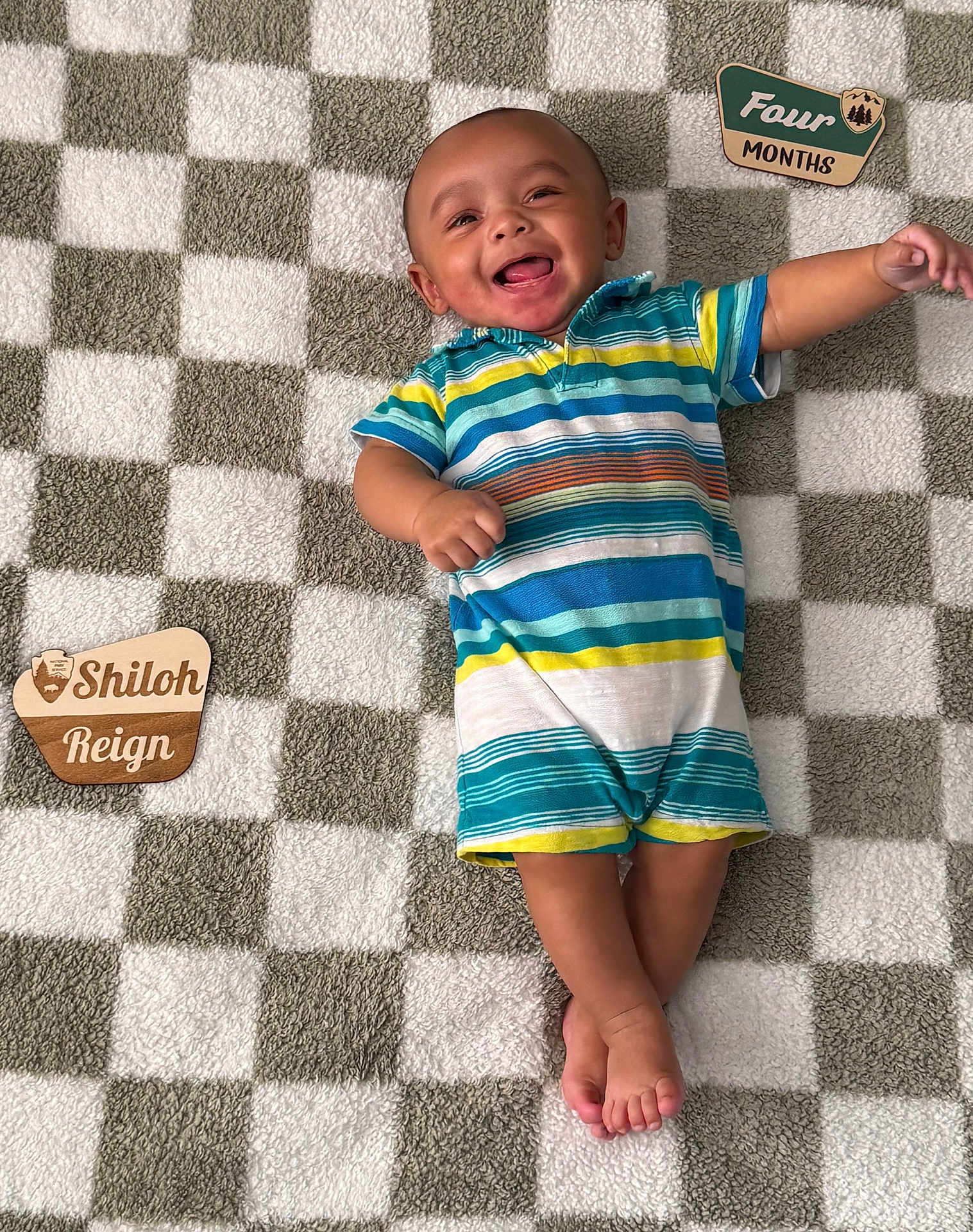 Shiloh is registered to the contest to win money with this photo: baby, child, smiling, striped_clothing, plush_blanket, checkered_pattern, wooden_plaques, infant, happy, lying_down, cute, portrait, indoors, feet, arms, head, person, colorful, playful, young