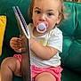 baby, book, casual_clothing, child, cozy, curly_hair, cushion, green_couch, indoors, pacifier, person, pink_shorts, playful, portrait, reading, relaxed, sitting, toddler, white_tshirt, wooden_wall