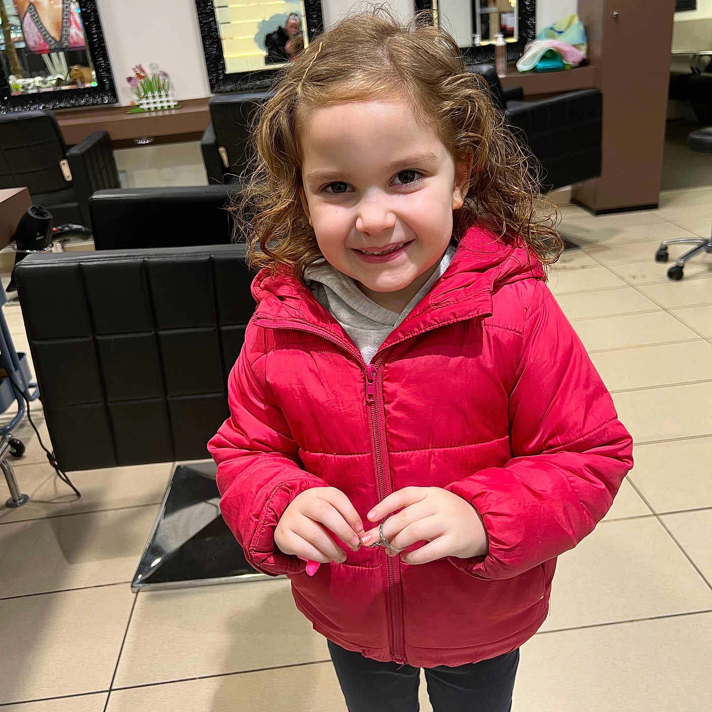 Alia a rejoint le concours — aidez-le/la à gagner de superbes lots ! bright_lighting, casual_clothing, chair, child, curly_hair, floor_tiles, girl, hair, hands, happy, indoor, mirror, person, portrait, red_jacket, reflection, salon, smile, standing, young