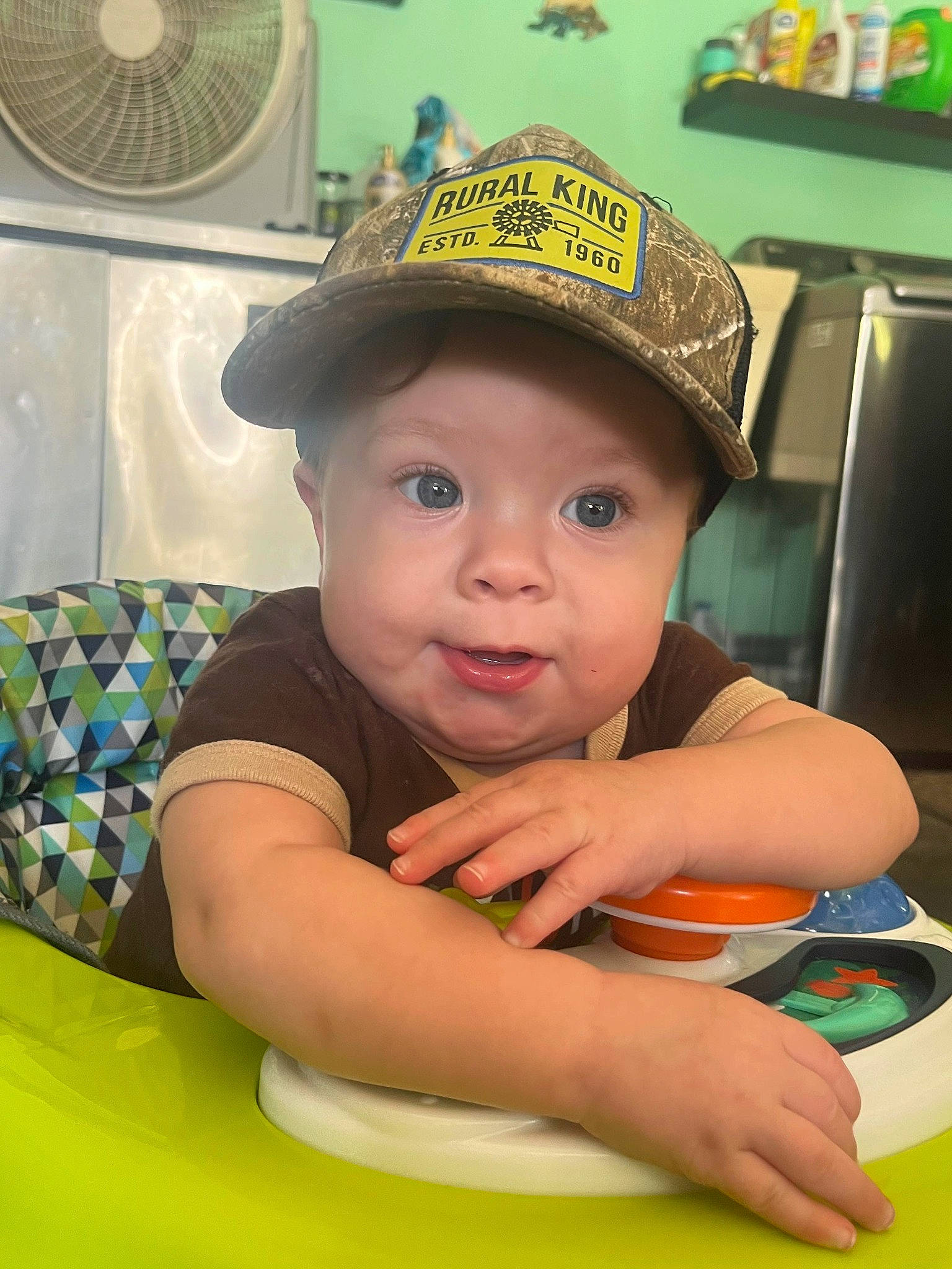Titus is registered to the contest to win money with this photo: baby, baseball_cap, cap, child, eye, fashion_accessory, fun, green, happy, hat, headwear, leisure, mechanical_fan, person, play, recreation, room, sitting, table, toddler
