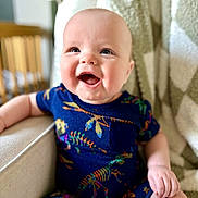 Owen is registered to the contest to win money with this photo: baby, smiling, child, infant, dinosaur_pattern, blue_clothing, sitting, chair, blanket, happy, cute, portrait, indoor, closeup, face, skin, person, young_child, cozy, comfort
