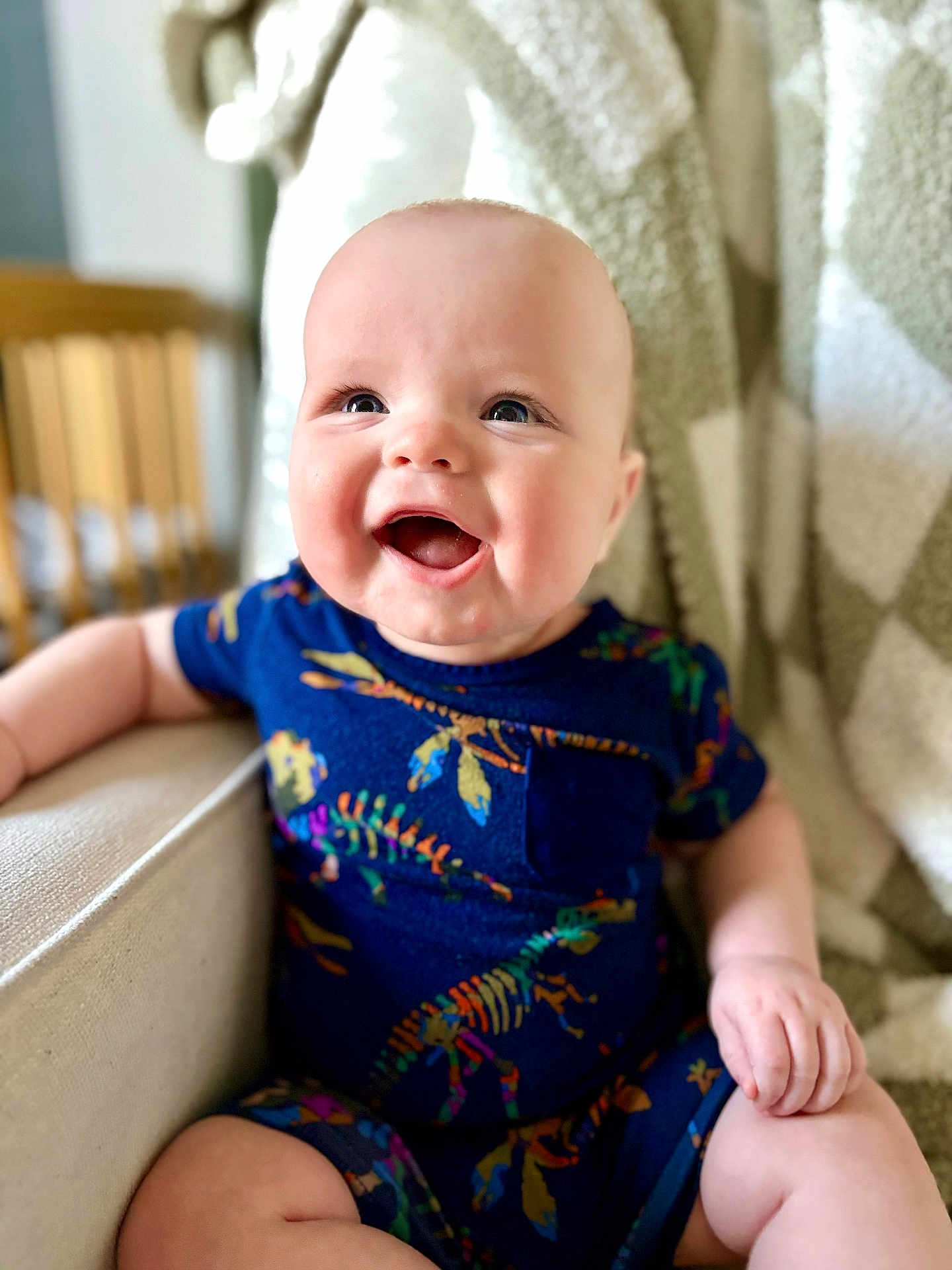 Owen is registered to the contest to win money with this photo: baby, smiling, child, infant, dinosaur_pattern, blue_clothing, sitting, chair, blanket, happy, cute, portrait, indoor, closeup, face, skin, person, young_child, cozy, comfort