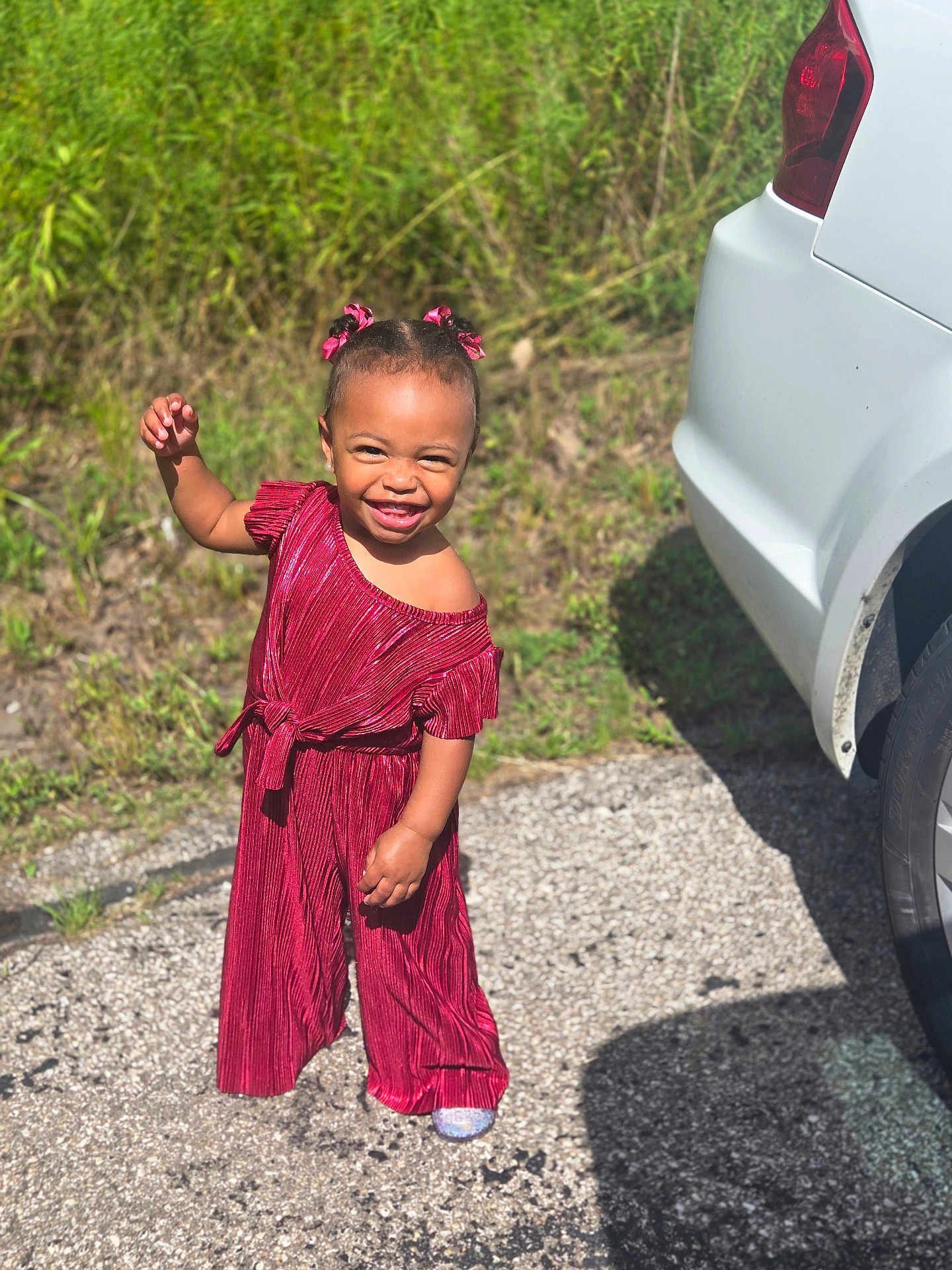 Symphony is registered to the contest to win money with this photo: child, toddler, smiling, red_clothing, outdoor, sunlight, grass, car, vehicle, pavement, happy, cute, portrait, person, fashion, hair_bows, standing, playful, daytime, nature