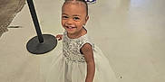 Symphony is registered to the contest to win money with this photo: toddler, child, dress, smile, indoor, floor, happy, person, hair_bow, white_dress, standing, footwear, legs, people, concrete_floor, wall, trash_can, black_pole, casual_clothing, joyful
