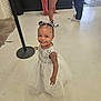 toddler, child, dress, smile, indoor, floor, happy, person, hair_bow, white_dress, standing, footwear, legs, people, concrete_floor, wall, trash_can, black_pole, casual_clothing, joyful