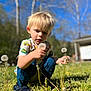child, toddler, dandelion, flower, grass, outdoor, nature, blue_sky, sunlight, curious, playing, greenery, young_child, casual_clothing, daylight, plants, grass_field, crouching, blond_hair, kid