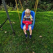 Logan is registered to the contest to win money with this photo: blonde_hair, blue_swing, book, casual_clothing, child, daylight, footwear, grass, greenery, happy, nature, outdoor, play, playground, reading, red_safety_bar, seated, swing, yellow_rope, young