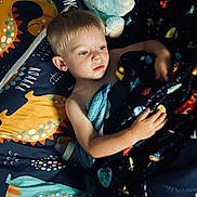 Logan is registered to the contest to win money with this photo: child, boy, bed, blanket, plush_toy, dinosaur, bedding, indoor, sleeping_area, light, face, hair, hand, blue, yellow, pattern, comfort, rest, portrait, young