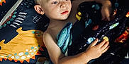 Logan is registered to the contest to win money with this photo: bed, bedding, blanket, blue, boy, child, comfort, dinosaur, face, hair, hand, indoor, light, pattern, plush_toy, portrait, rest, sleeping_area, yellow, young