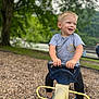 child, smiling, playground, seesaw, outdoor, tree, woodchips, casual_clothing, blue_shoes, greenery, happy, daylight, park, short_sleeves, pants, playtime, young_child, nature, fun, person