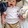 baby, bassinet, beige_shorts, child, child_clothing, comfort, cozy, cushion, cute, fabric, indoor, infant, plush_toys, portrait, sleeping_area, small_hands, small_legs, soft_toys, surprised_expression, white_polo_shirt