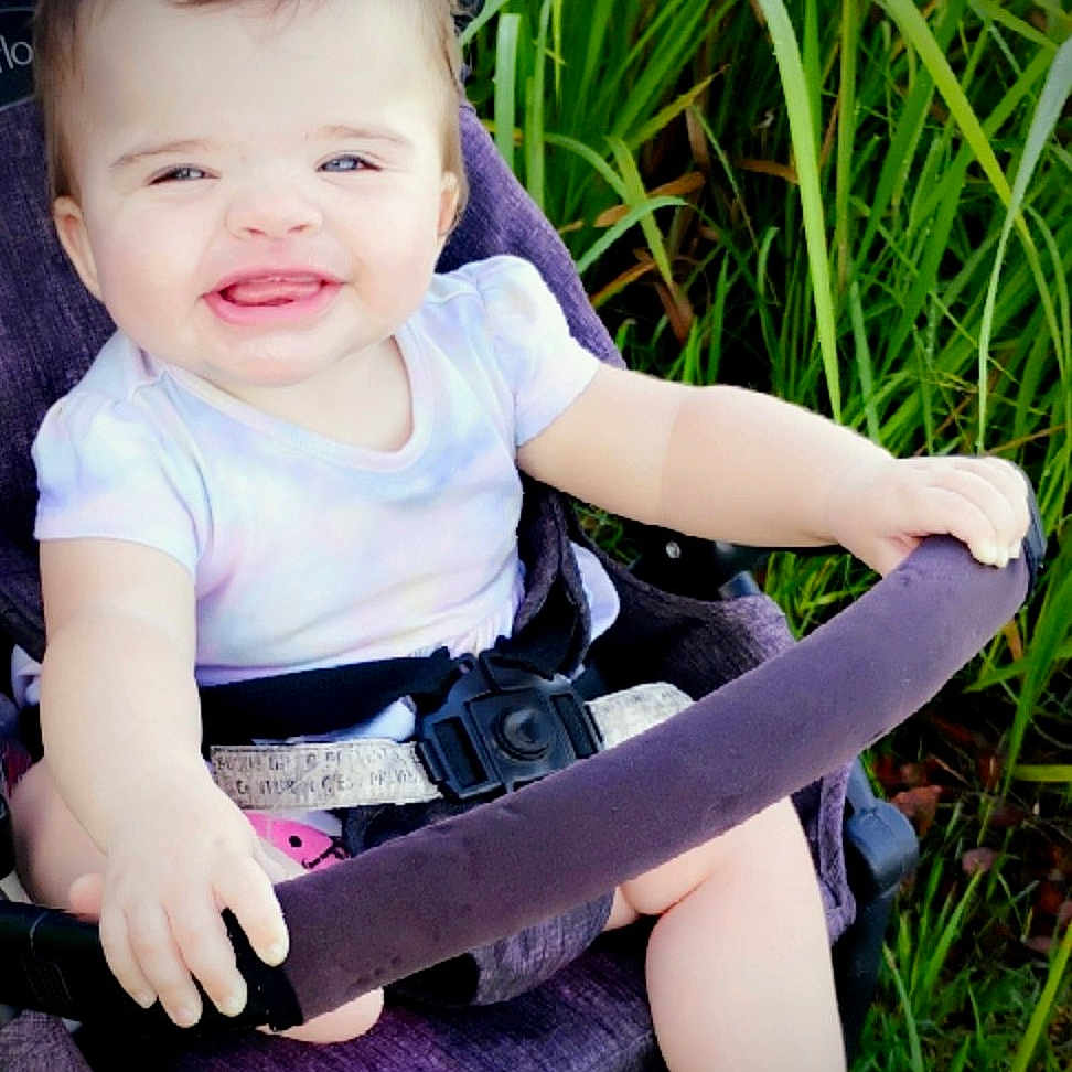 Adelina-Jo is registered to the contest to win money with this photo: baby, child, clothing, cute, daylight, foot, grass, green, hand, happy, infant, nature, outdoor, person, portrait, purple, seatbelt, sitting, smiling, stroller