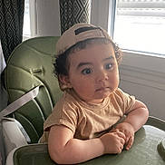 Yanis a rejoint le concours — aidez-le/la à gagner de superbes lots ! toddler, child, high_chair, cap, curly_hair, beige_shirt, green, window, curtains, indoor, natural_light, face, portrait, expression, person, furniture, seat, young_child, hands, casual