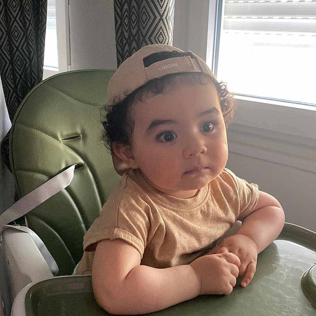 Yanis a rejoint le concours — aidez-le/la à gagner de superbes lots ! toddler, child, high_chair, cap, curly_hair, beige_shirt, green, window, curtains, indoor, natural_light, face, portrait, expression, person, furniture, seat, young_child, hands, casual