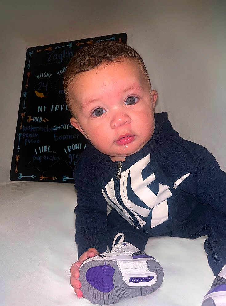Zaylin is registered to the contest to win money with this photo: baby, child, sitting, hoodie, sneaker, shoe, white_surface, blackboard, handwriting, indoor, curious, young_child, clothing, footwear, portrait, face, casual, purple, bright_eyes, person
