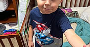 Zachariah is registered to the contest to win money with this photo: child, boy, indoor, bedroom, crib, blanket, clothing, blue_shirt, car_graphic, carpet, wooden_furniture, toy, smiling, person, footwear, shorts, messy_room, curtain, wall, expression