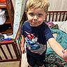 child, boy, indoor, bedroom, crib, blanket, clothing, blue_shirt, car_graphic, carpet, wooden_furniture, toy, smiling, person, footwear, shorts, messy_room, curtain, wall, expression