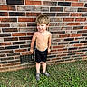child, boy, shirtless, shorts, sneakers, brick_wall, grass, outdoor, smiling, face, standing, dirty_face, playful, casual, daylight, young, person, nature, summer, portrait