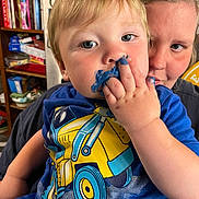 Wesley is registered to the contest to win money with this photo: toddler, child, adult, face, blue_paint, hand, shirt, toy_truck_print, person, indoor, bookshelf, furniture, curly_hair, expression, skin, closeup, holding, smudge, blue_clothing, background