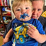 Wesley is registered to the contest to win money with this photo: adult, blue_paint, bookshelf, child, closeup, clothing, cookbook, expression, face_paint, family, hand, holding, indoor, kitchen, messy, person, portrait, sitting, tissue, toddler