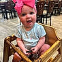Claire is registered to the contest to win money with this photo: baby, child, clothing, cute, expression, flooring, furniture, happy, headwear, high_chair, holding, indoor, infant, person, pink_bow, restaurant, seat, smiling, table, wooden_chair