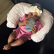 Ava joined the competition — help win amazing prizes! baby, infant, bottle, headband, pink_clothing, cushion, sofa, indoor, child, cute, relaxing, looking_at_camera, comfortable, small_hands, foot, dark_furniture, portrait, human, toddler, feeding